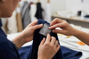 Close-up das mãos de uma pessoa tocando uma etiqueta cinza em relevo fixada em uma peça de roupa azul-marinho. A etiqueta exibe formas geométricas táteis como um círculo, um triângulo, um quadrado e uma grade de linhas cruzadas, representando o sistema de identificação para deficientes visuais em um ateliê de costura.